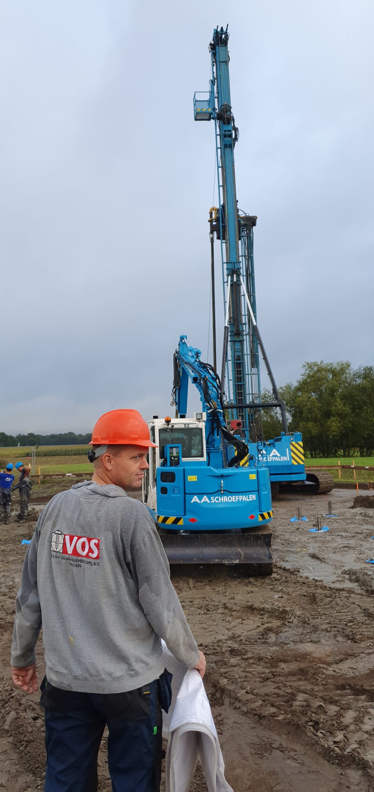 Bouwvakker met oranje helm voor blauwe heipaalmachine op modderige bouwplaats.