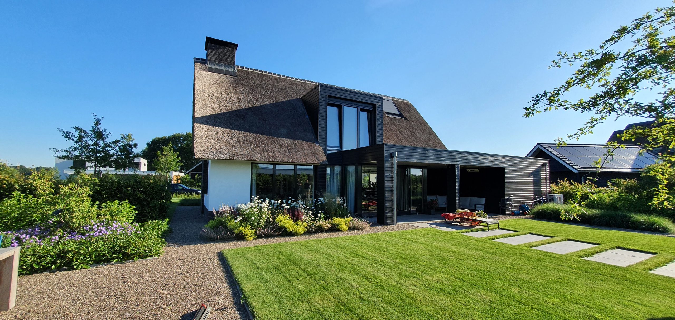 Moderne woning met rieten dak, grote ramen en groene tuin onder blauwe lucht.