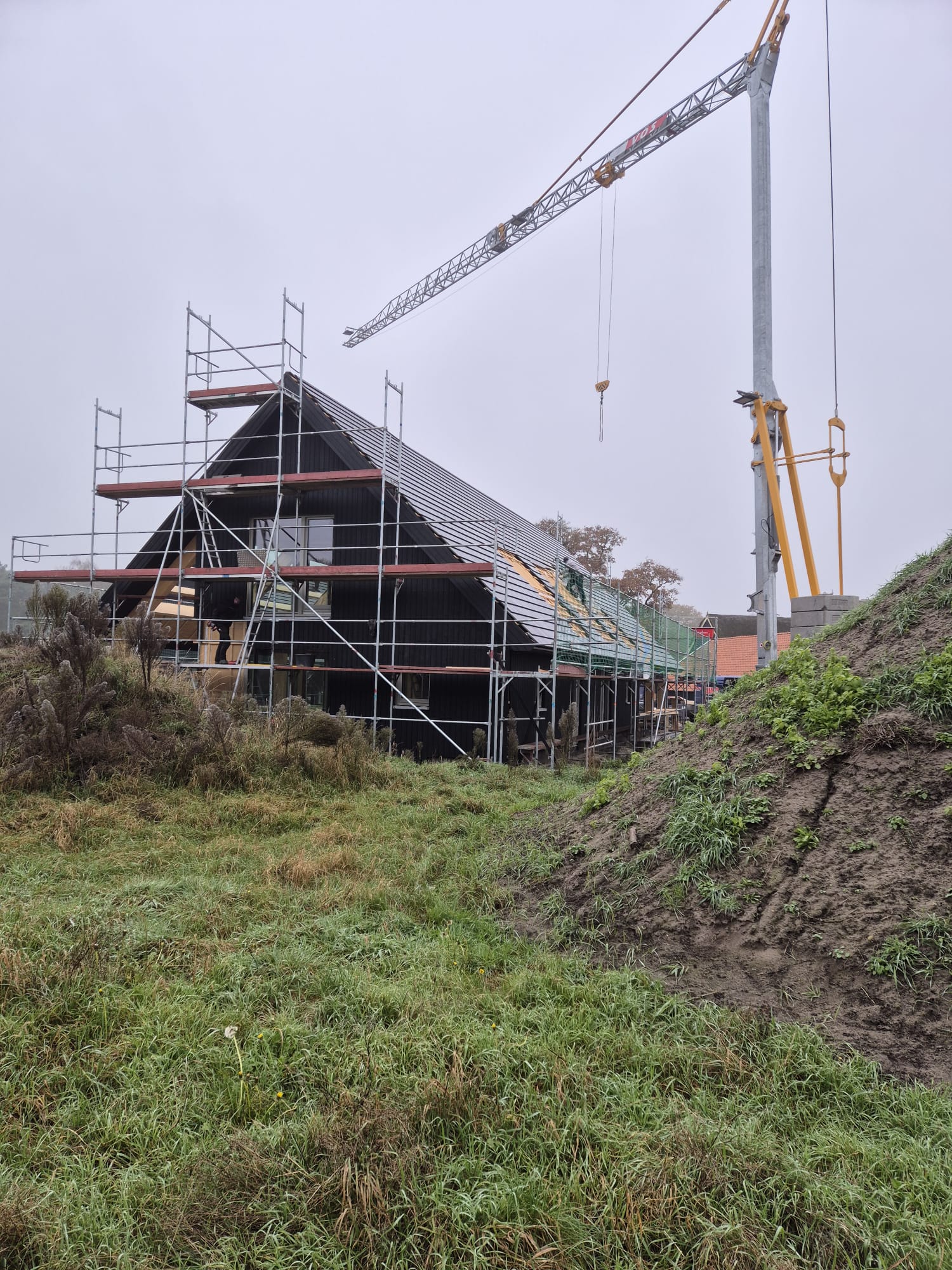 Zwart houten huis in aanbouw met steigers en kraan op een natte weide.