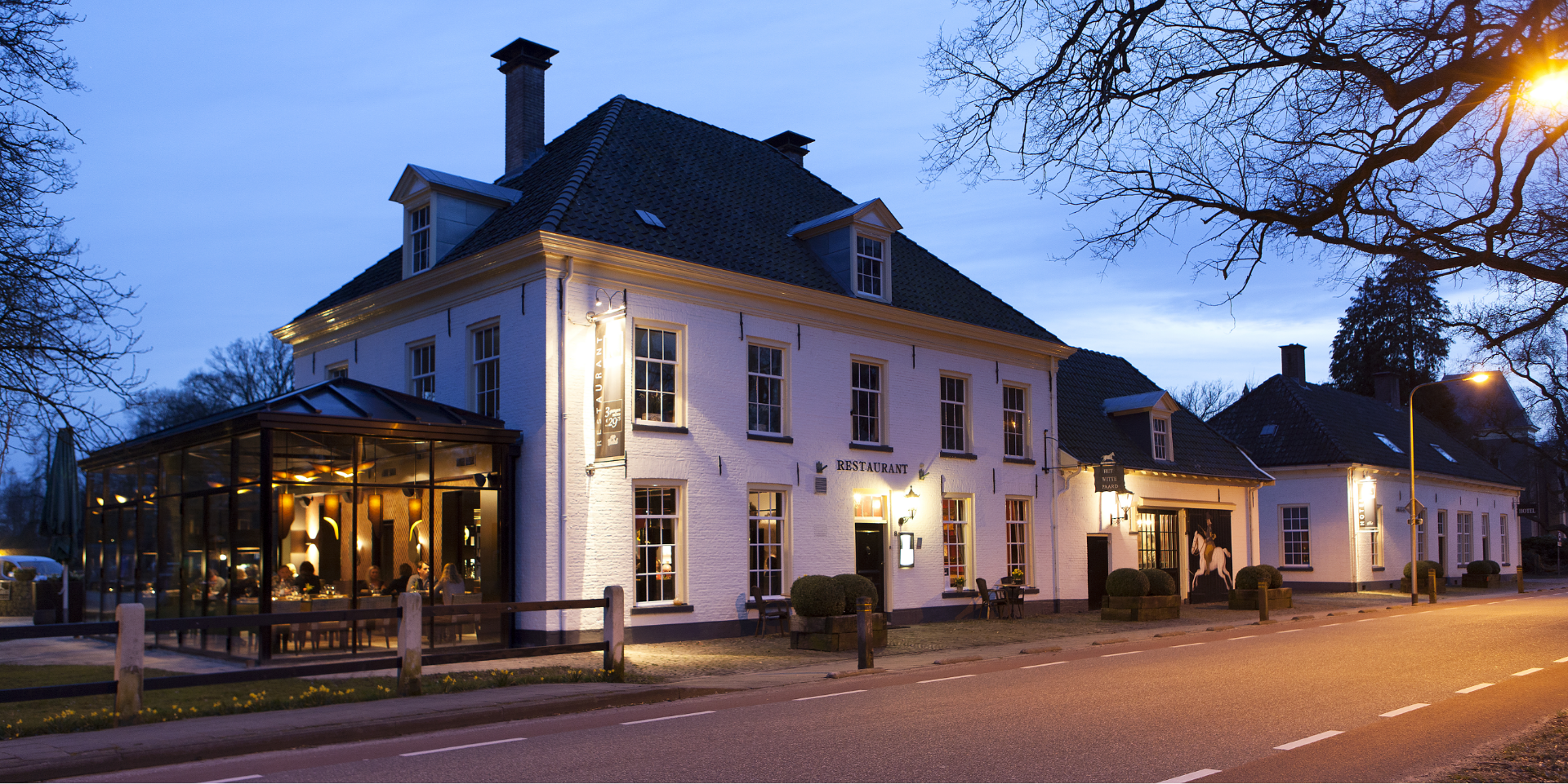 Historisch wit restaurant met glazen serre, sfeervol verlicht bij avond.